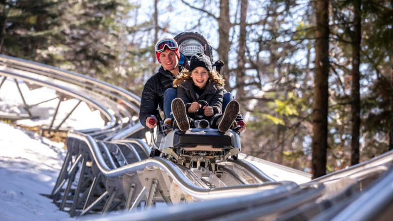 Alpine rollercoaster on a snowy hill, two people riding a coaster car on the track.