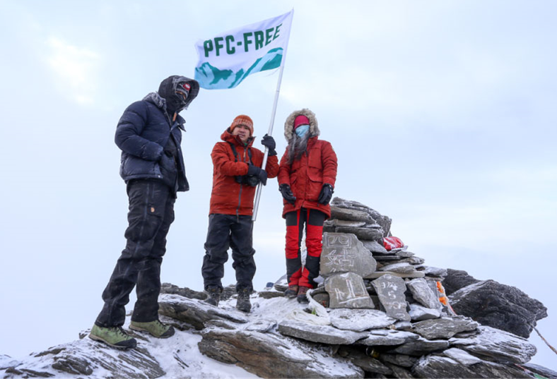 Three people in snow wear holding a flag a top a rocky top with a dusting of snow covering it.