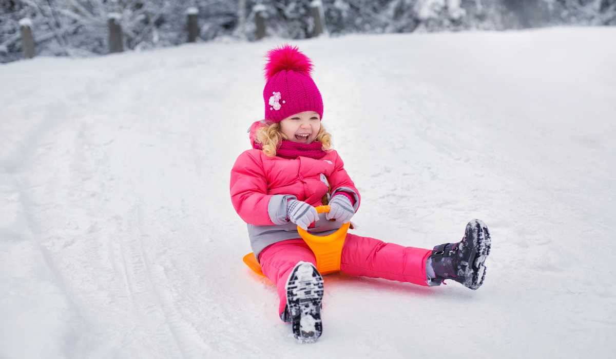 small girl in pink bobble hat riding on a toboggan down a snowy slope.