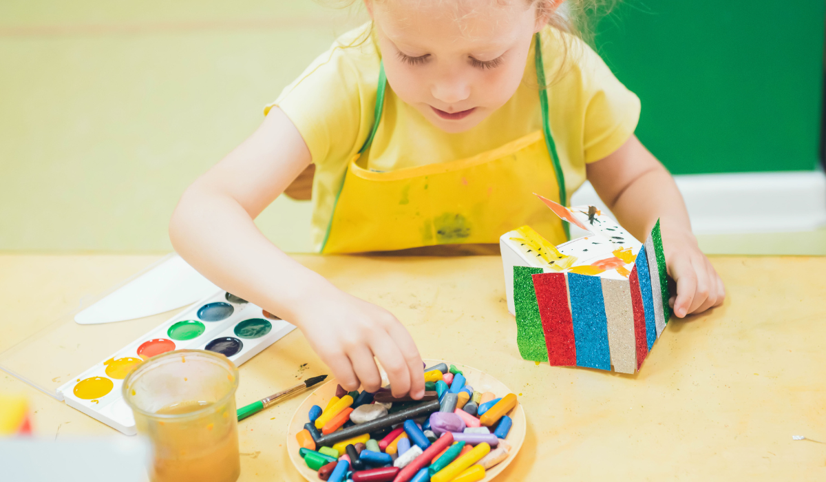 young girl in yellow tshirt is crafting on a table, She is creating a mini-hoiuse with colourful roof and using paints and crayons to decorate it.