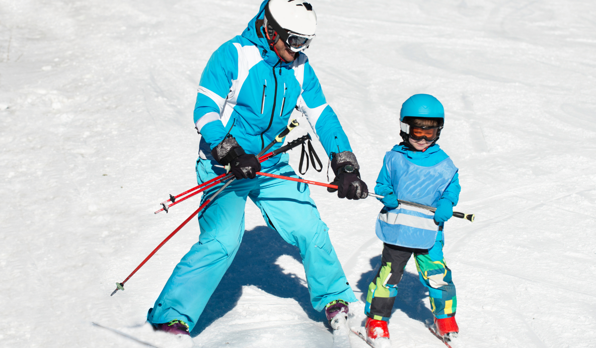 An adult is holding a pole, guiding a small child down a small nursery ski slope.
