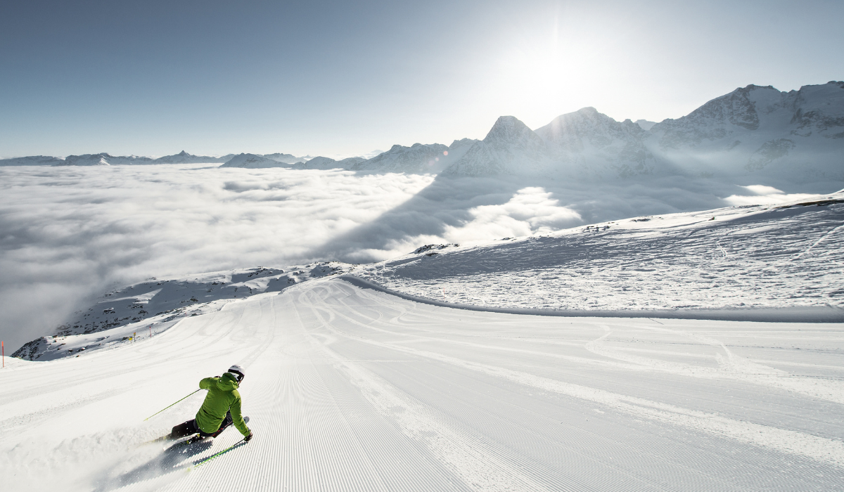 Above clouds view of a piste with sunny sky and a skiier carving down the cordoroy piste.